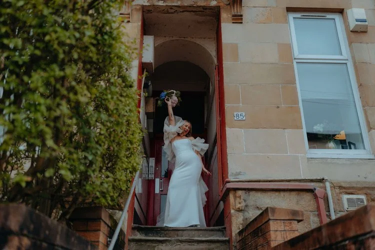 A bride stands proudly in the doorway of a city townhouse, lifting her bouquet into the air with confidence and joy, captured with bold creativity by F.D Young Photography. The textured stone entrance, dramatic lighting and stylish gown create a dist