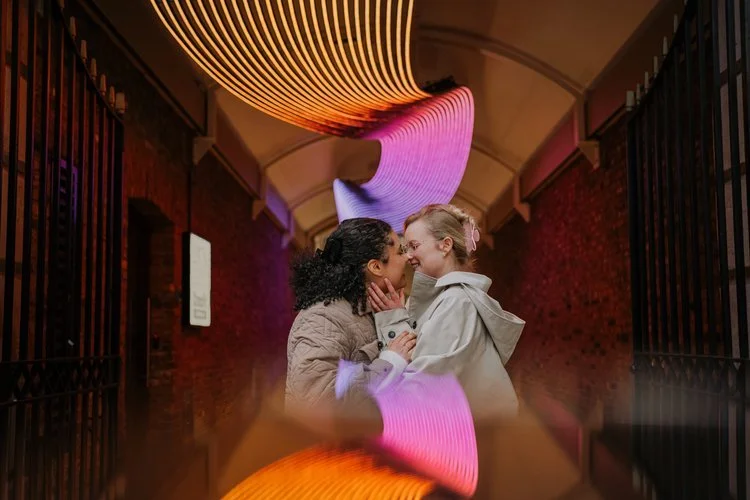 A couple share a joyful intimate moment beneath vibrant neon-style light trails in an urban archway, captured by Joshua Humphrey Photography. The colourful reflections and warm candid connection create a modern artistic portrait perfect for contempor