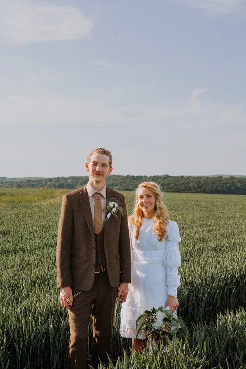 A couple stand hand in hand in a sunlit countryside field, captured by Joshua Humphrey Photography. The groom wears a rustic brown suit while the bride holds a natural bouquet and smiles warmly, creating a relaxed rural wedding portrait with soft gol