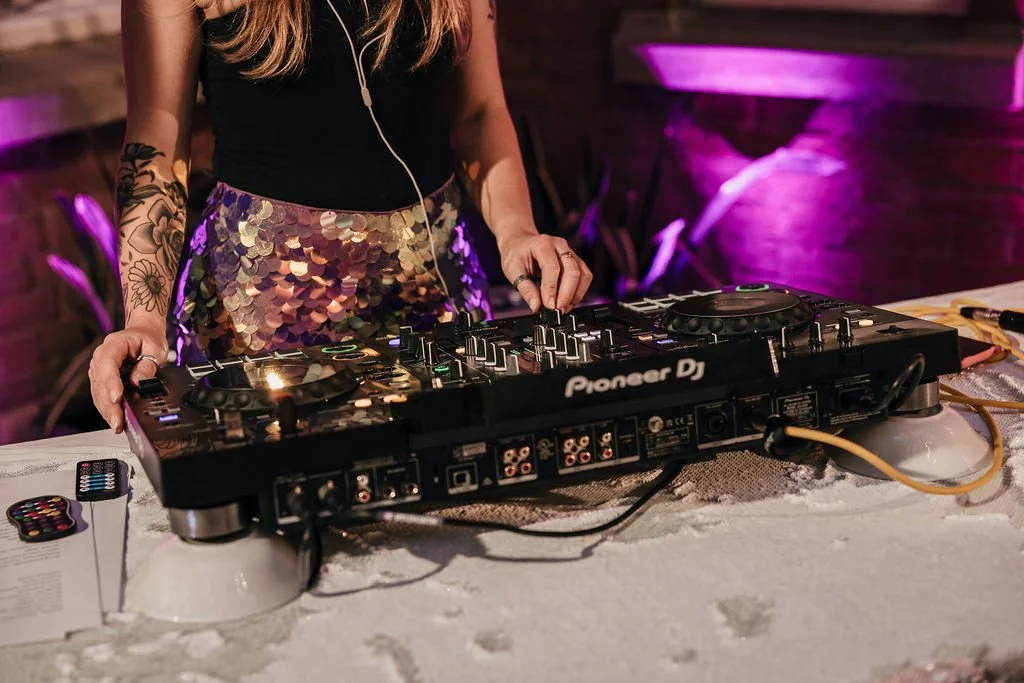 Close-up of a Rave the Date wedding DJ mixing tracks on a Pioneer DJ controller, captured by Jules Barron Photography. The DJ’s sequin skirt and tattooed arm add a touch of sparkle and individuality, while the soft pink and purple lighting sets a fun