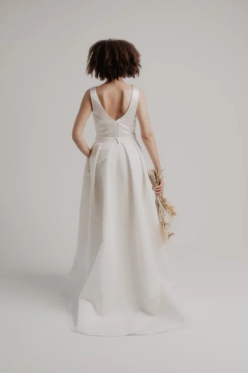 Sarah Barker elegant plunge back ballgown wedding dress in ivory satin with a full skirt and button detail, photographed in a minimalist studio setting with the bride holding a dried flower bouquet.