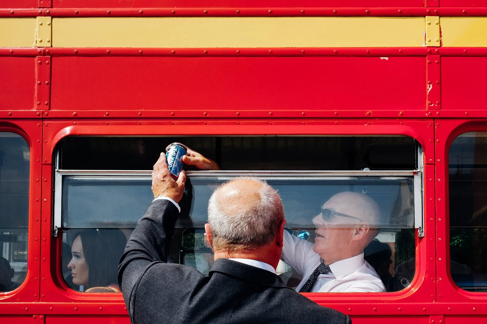 A man in a suit stands outside a bright red vintage bus, passing a can through the open window to another man seated inside who is wearing sunglasses and a white shirt. A woman seated further along the bus looks out of her window while sunlight highl