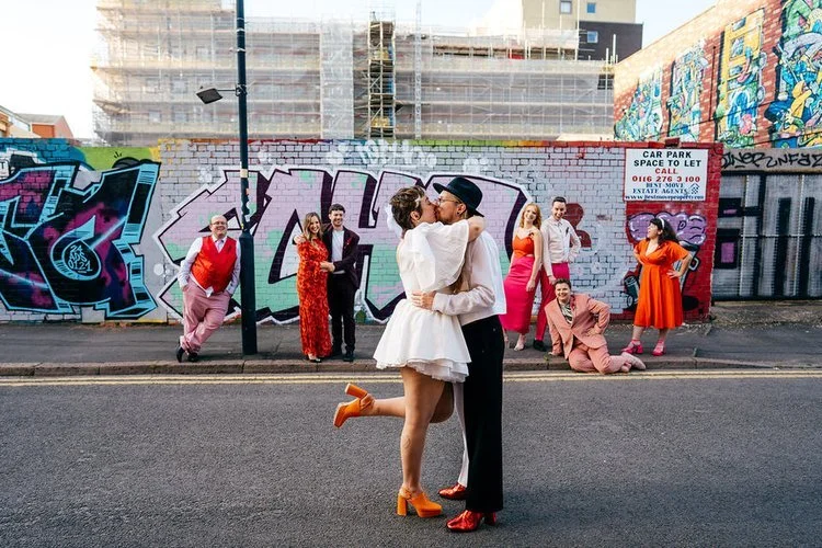 A newly married couple share a kiss in front of colourful graffiti walls while their wedding party cheer, pose and celebrate around them, captured with bold energy by Jordanna Marston Photography. The bright outfits, urban street art and playful reac