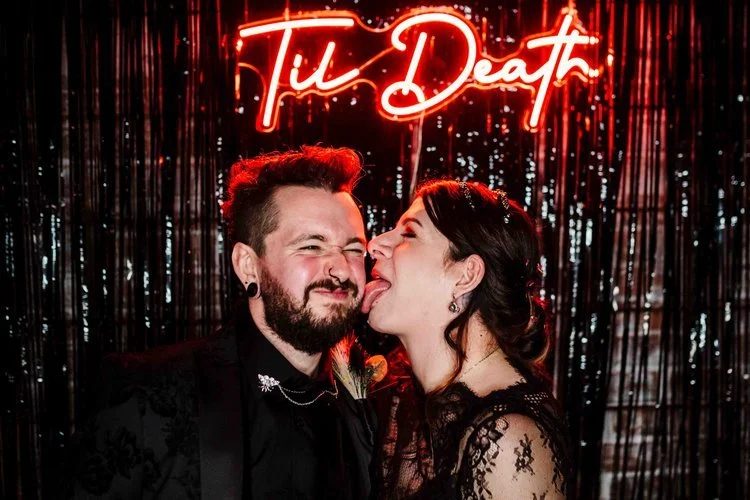 A bold and energetic wedding moment captured by Amy Faith Photography, showing the newlyweds celebrating on the steps with a lively brass band and an ecstatic crowd behind them. The bride, still in her veil and holding a vibrant bouquet, beams with j