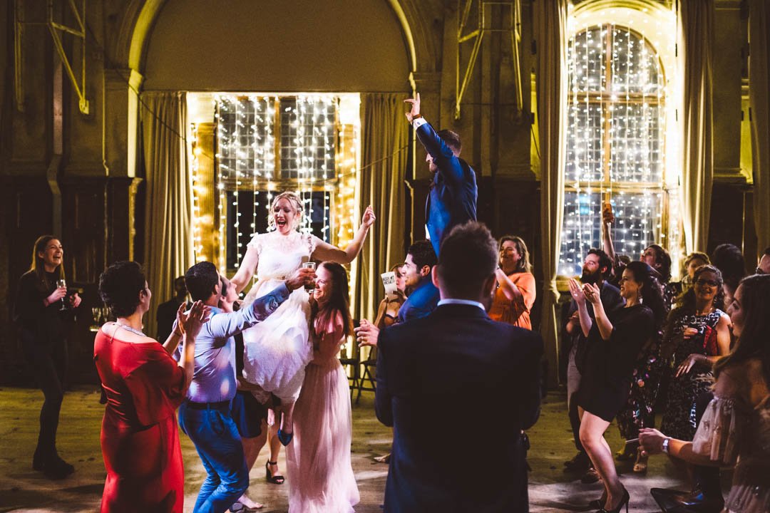A high-energy wedding dancefloor moment captured by Barbara K Photography, featuring the bride being lifted joyfully into the air by friends as guests cheer and celebrate around her. Set inside a grand London venue with dramatic high ceilings and spa