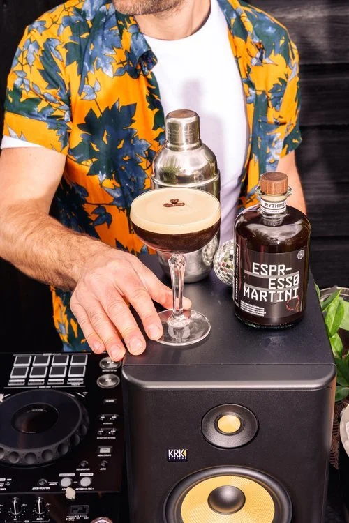 A man wearing a vibrant floral shirt stands behind DJ equipment, holding a freshly shaken Espresso Martini beside a Rythmik Cocktails bottle of the same drink. A cocktail shaker and small disco ball add to the lively setup, blending music and mixolog