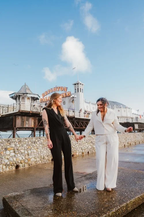 A couple walk hand in hand along the seafront beside the iconic Brighton Palace Pier, smiling at one another as the sunlight reflects off the wet pavement. Their stylish outfits - a sleek black jumpsuit and a chic white suit - give the moment a moder