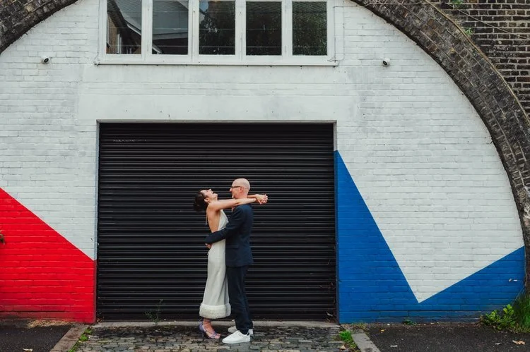 A couple embrace and laugh together in front of a bold, geometric mural painted on a white brick archway, blending vibrant red and blue shapes with the urban texture of the city. The bride’s modern, minimalist outfit and the groom’s relaxed suit give