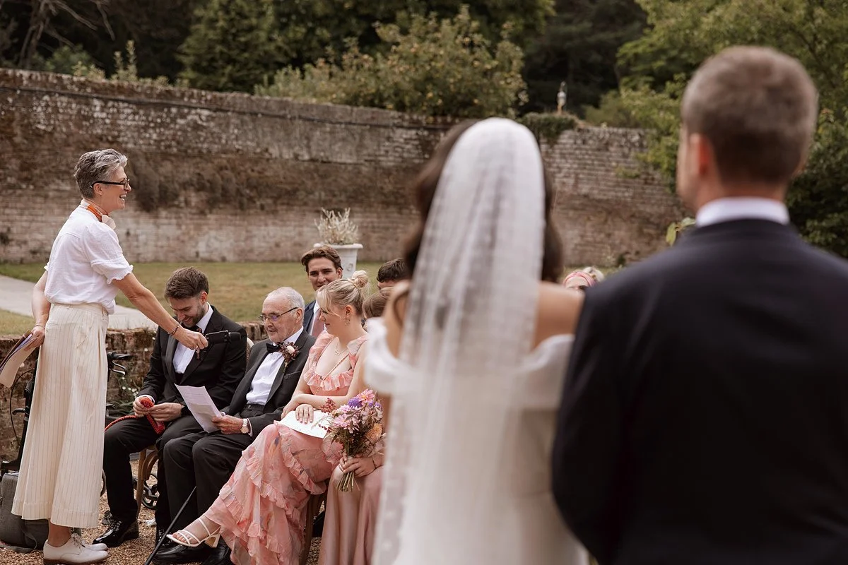Arabella stands to one side of the seated guests, leaning in slightly as she gestures toward a person holding a piece of paper, while the couple are visible in the foreground, out of focus. The scene is set outdoors against a stone wall and greenery,