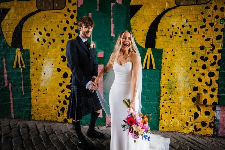 A joyful bride and groom pose against a vibrant yellow and green street-art mural, creating a fun and modern urban wedding portrait. The bride, wearing a sleek white dress and holding a bright bouquet of colourful flowers, laughs while the groom in a