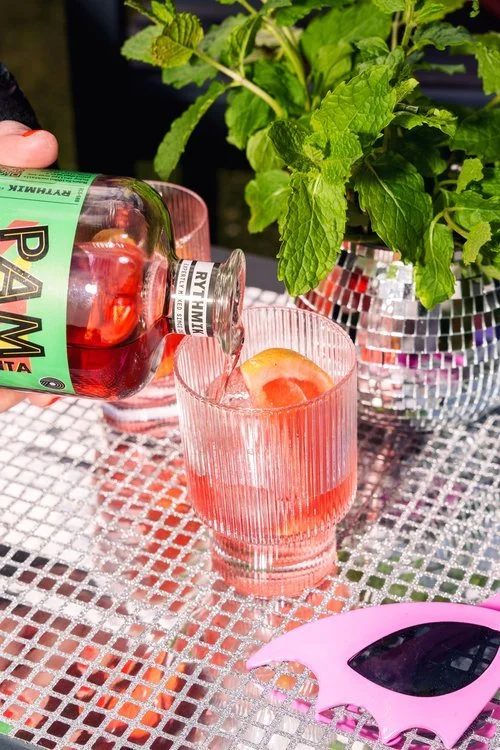 A bottle of Rythmik’s Ramalama Margarita is being poured into a textured glass over ice, garnished with a slice of citrus. The drink sits on a reflective silver table adorned with a disco ball planter and fresh mint, adding a lively and stylish touch