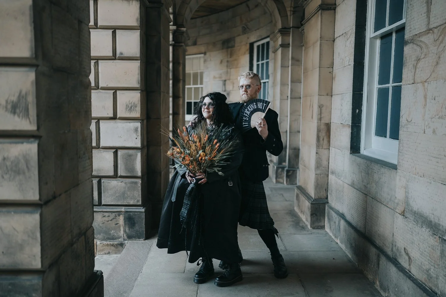 A bold, alternative wedding portrait in Glasgow showing a couple dressed in black beneath historic stone arches, with the bride holding a dried autumnal bouquet and the groom in a kilt holding a playful statement fan, captured in a moody, documentary