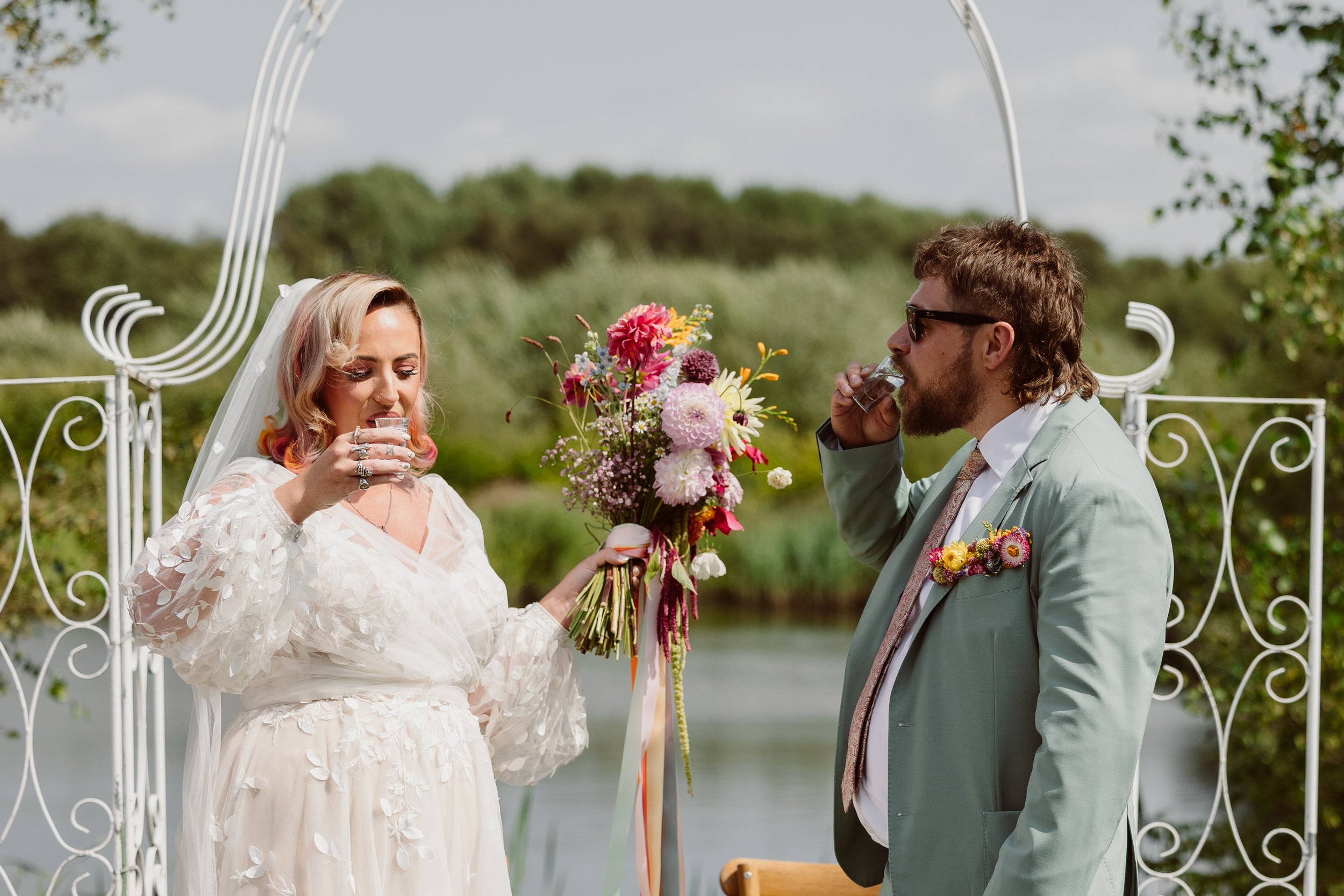 A relaxed and heartfelt ceremony moment captured by Joy Louise Photography, showing a bride and groom sharing a drink together beside a lakeside backdrop. The bride holds a vibrant bouquet filled with colourful summer flowers and ribbon streamers whi