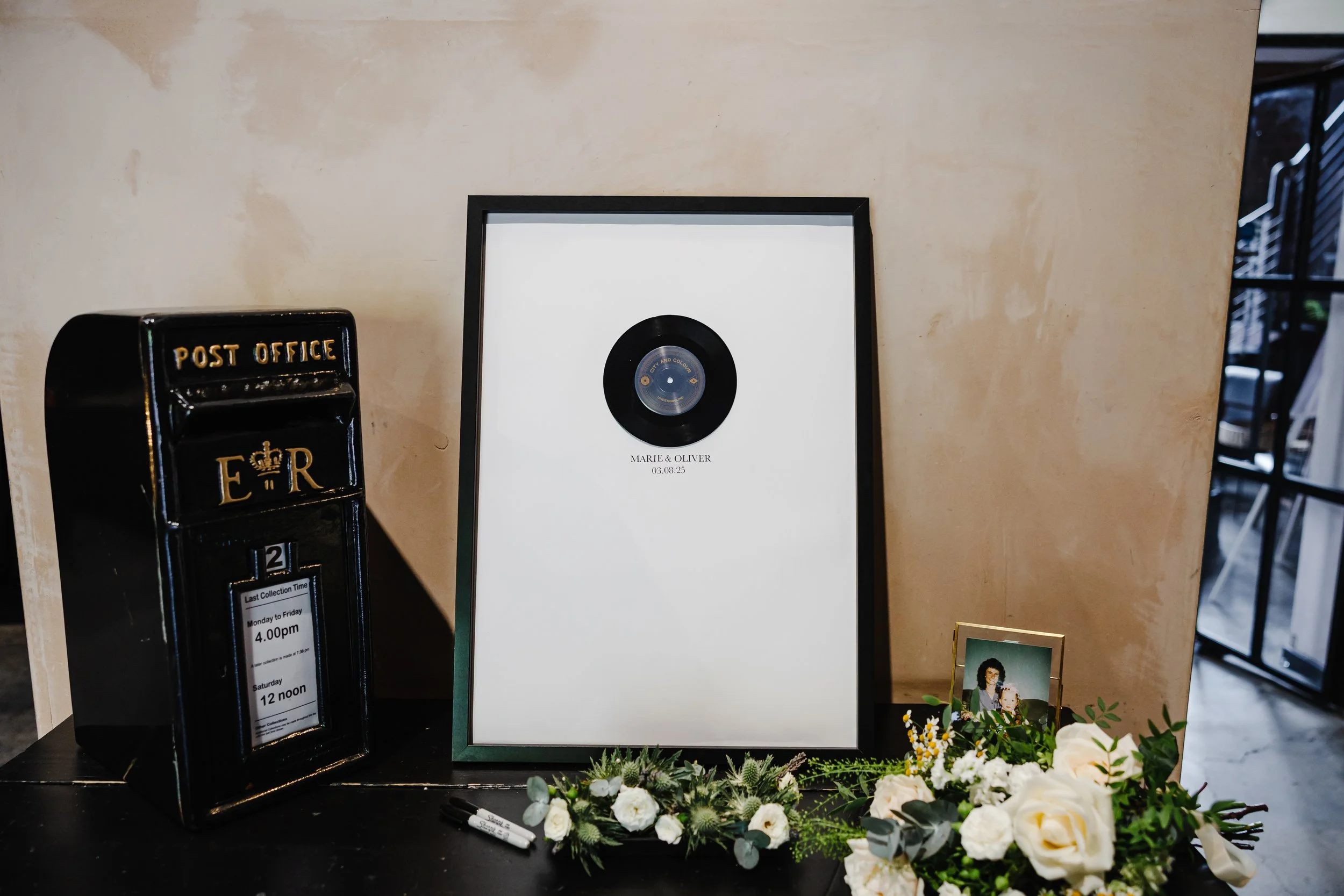 A stylish wedding memory table features a framed personalised vinyl record print for Marie & Oliver, set beside a classic black post box. White and green florals, guestbook pens, and a small framed photograph create a meaningful tribute display.