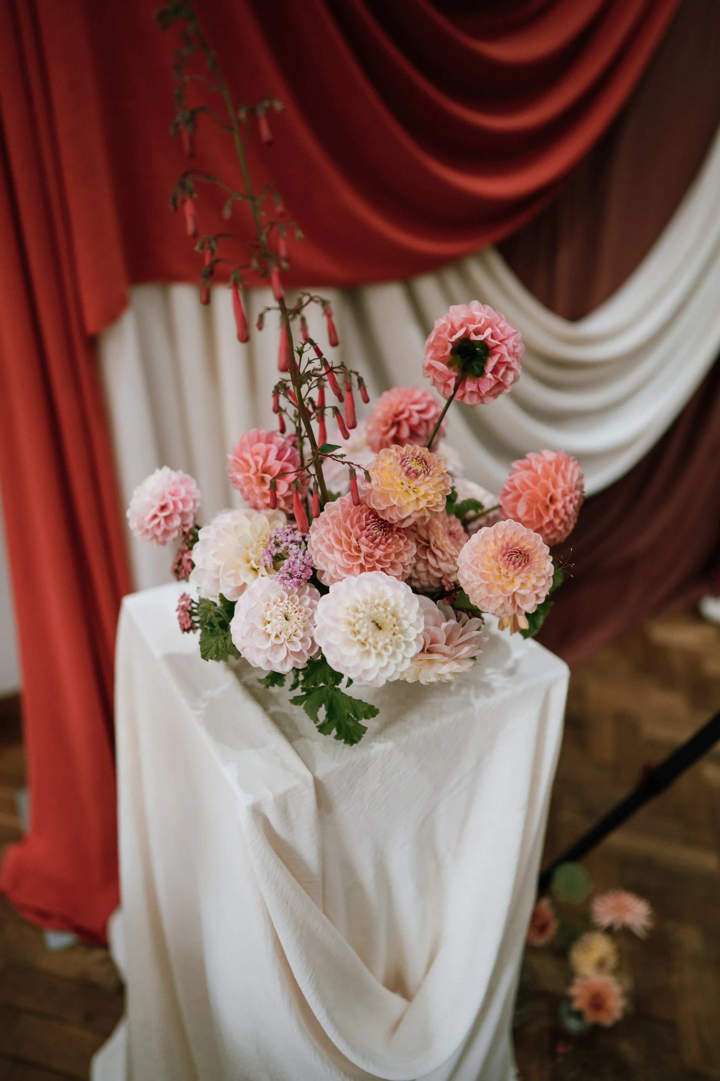Orange Tree Weddings styling and décor featuring soft pink and blush floral arrangements on draped white plinths, set against layered terracotta and cream fabric backdrops for elegant, editorial wedding styling.