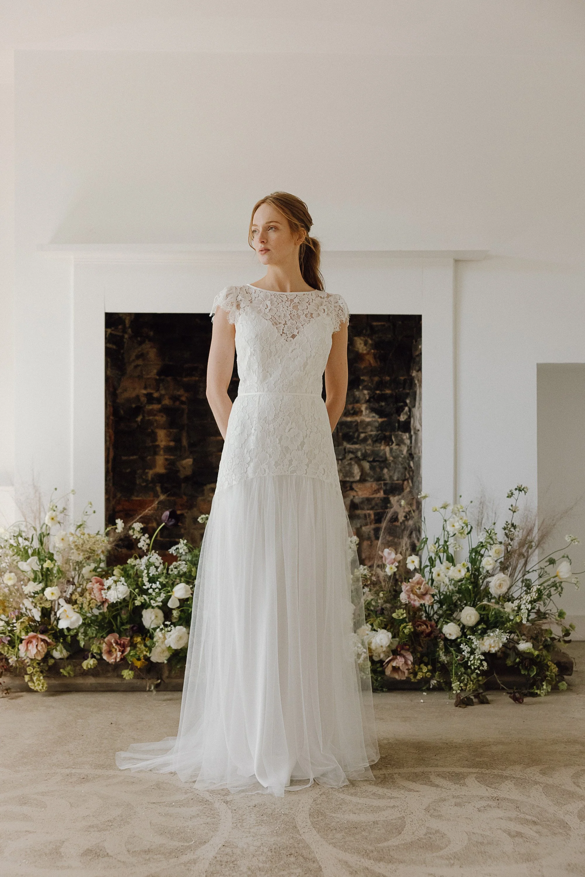 The bride stands softly lit in an elegant room, wearing a lace bodice gown with delicate cap sleeves and a sheer tulle skirt that falls lightly to the floor, while a bed of pastel flowers arranged around the fireplace adds a romantic garden-inspired 