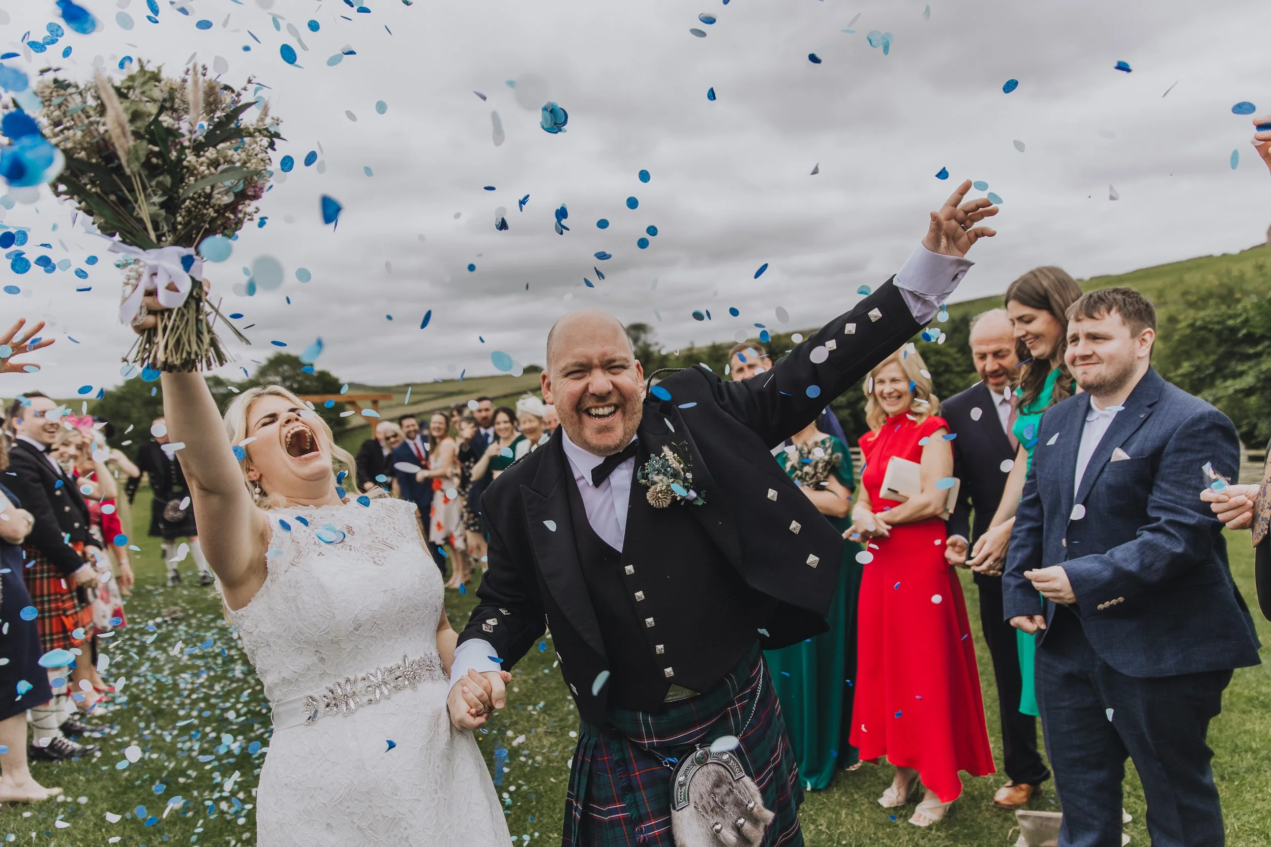 A high energy confetti moment captured by YU Photography, showing the newly married couple erupting with pure joy as blue and white confetti rains down around them. The bride throws her bouquet triumphantly into the air while laughing with unfiltered