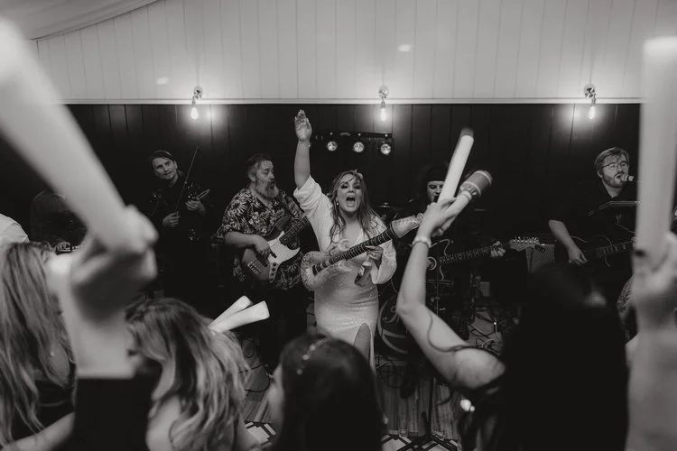 A bride takes centre stage with a guitar in hand, singing to a cheering crowd as the live band performs behind her, captured with powerful energy and atmosphere by F.D Young Photography. Guests raise glow sticks in celebration while the monochrome to