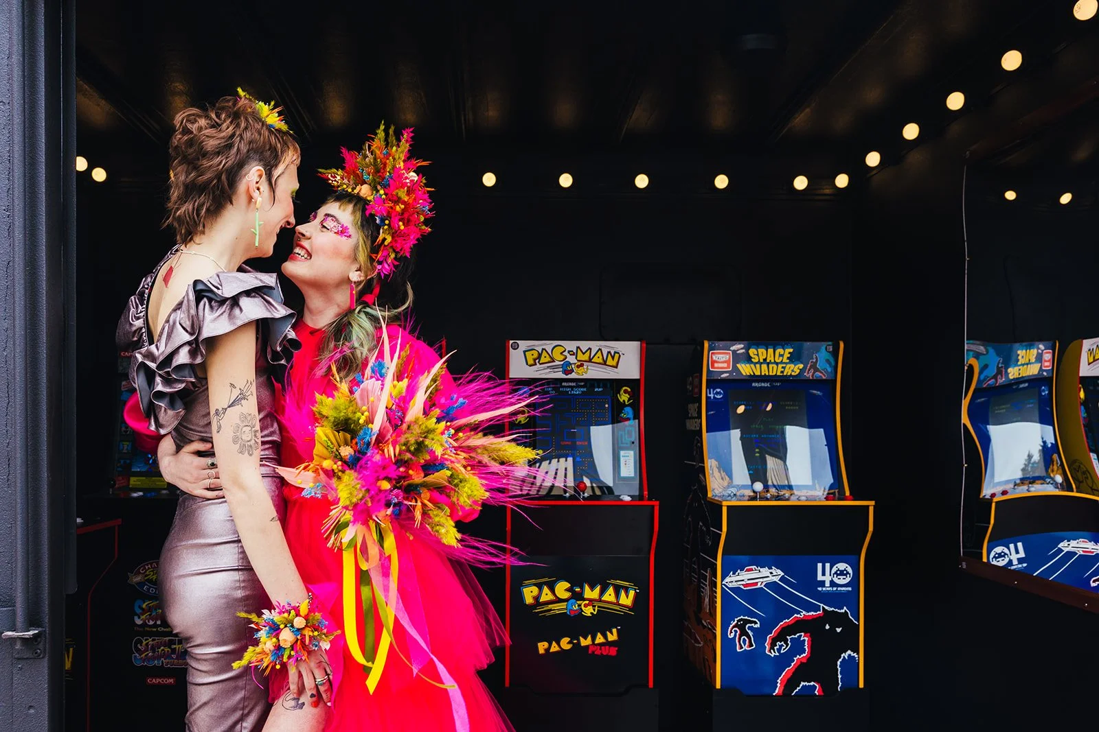 Two people share a joyful, electric moment surrounded by retro arcade machines, their vibrant dried flower pieces exploding with neon pink, yellow, blue and green that echo the playful energy of their outfits while the dramatic lighting and saturated