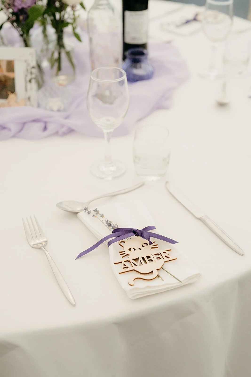 A wooden, cat shaped place setting on a white wedding table. It has the name 'Amber' and there is a purple bow wrapped around it.