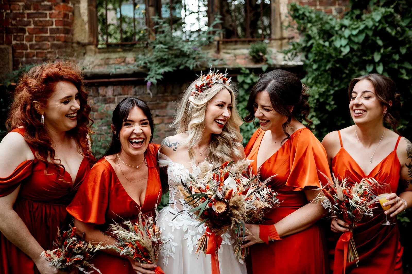 A joyful group of women in vibrant red dresses surrounds a smiling bride whose dried flower bouquet blends warm rust, cream and apricot tones, with feathery grasses and textured stems echoing the bold headpiece in her hair as laughter and closeness f