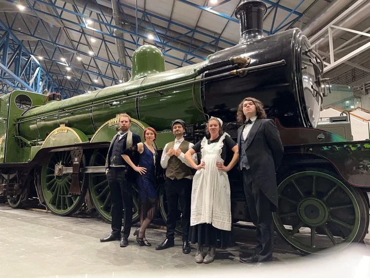 A group of actors in Victorian clothes, standing in front of a steam train.