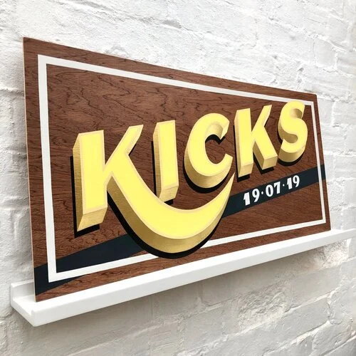 A bespoke wooden sign by Esther of Sylvie Signs featuring bold 3D yellow lettering spelling “KICKS” with the date “19.07.19” in navy blue. The hand-painted design uses playful vintage typography with layered shadows on a rich wood grain background fr