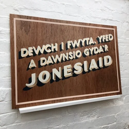 A custom Welsh-language wooden sign hand-painted by Esther of Sylvie Signs reads “Dewch i fwyta, yfed a dawnsio gyda’r Jonesiaid,” meaning “Come eat, drink, and dance with the Joneses.” The lettering is painted in a bold cream and black 3D style, fra