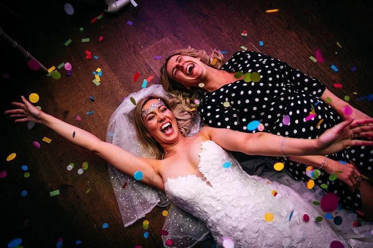 A bride and guest lie on the dancefloor laughing and throwing their arms in the air as colourful confetti falls around them, captured with energy and joy by Jordanna Marston Photography. The bold colours, expressive reactions and party atmosphere cre