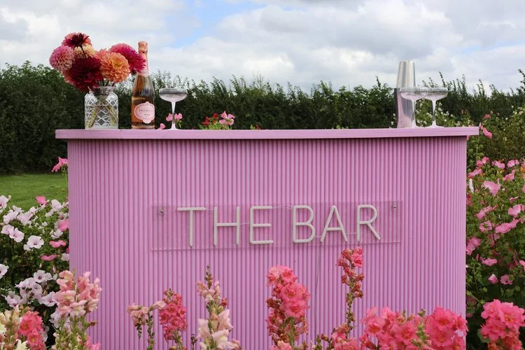 A vibrant pink outdoor bar setup by The Pink Drinks Van, surrounded by blooming flowers and countryside greenery. The bar features elegant signage reading “THE BAR,” with bottles of sparkling rosé, cocktail glasses, and fresh dahlias creating a chic 