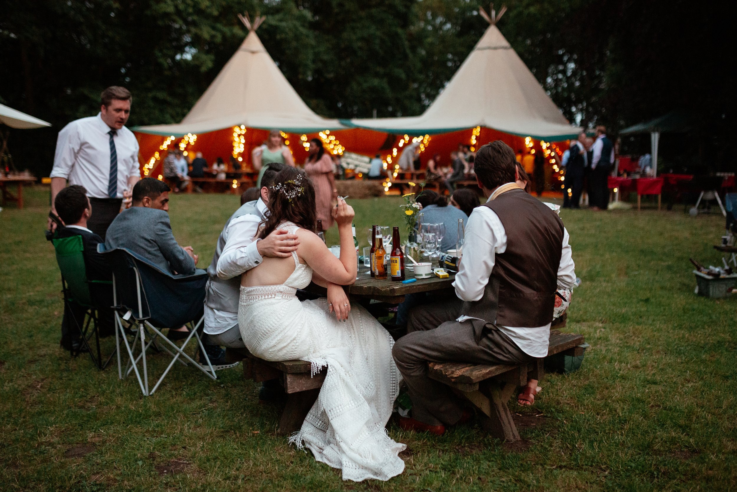 A relaxed and atmospheric evening wedding scene captured by Laura Wilson Photography, showing guests gathered around picnic tables outside a tipi reception. The bride sits with her partner and friends, leaning in for a quiet moment while warm string 