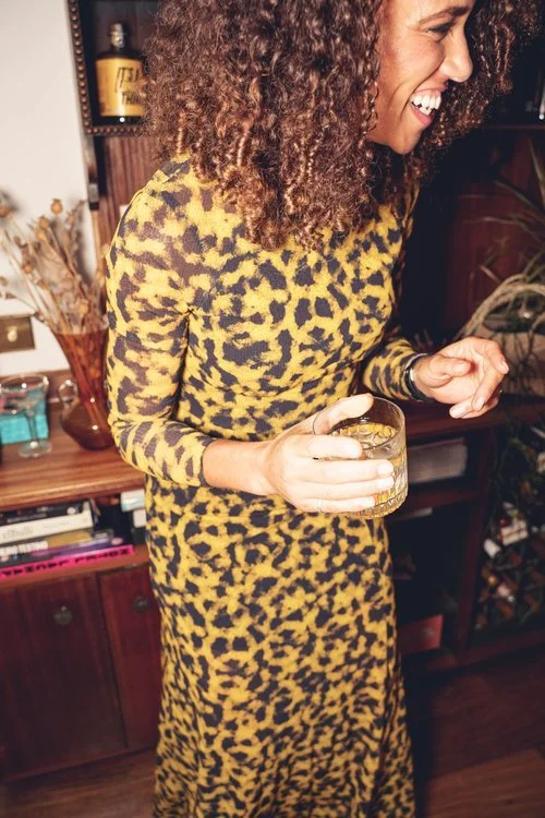A woman in a bold yellow and black leopard-print dress laughs while holding a glass of Rythmik’s handcrafted cocktail in a cosy indoor setting. The drink’s golden hue glows in the warm light, perfectly complementing the relaxed, sociable atmosphere. 