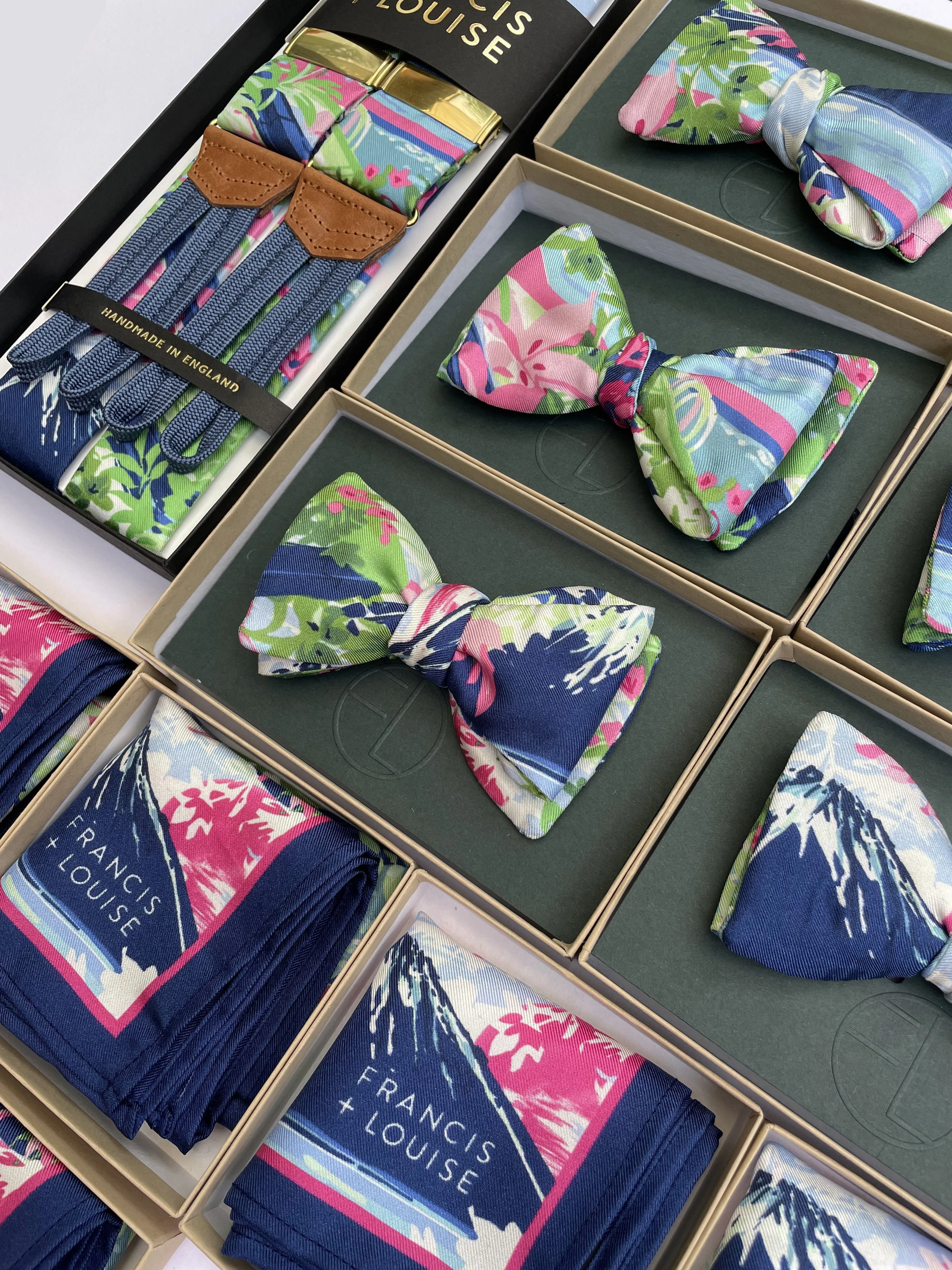 An arrangement of Francis and Louise accessories neatly displayed in individual boxes, including patterned bow ties, pocket squares and a pair of suspenders. Each item features a vibrant multicoloured design with shades of blue, pink and green, incor