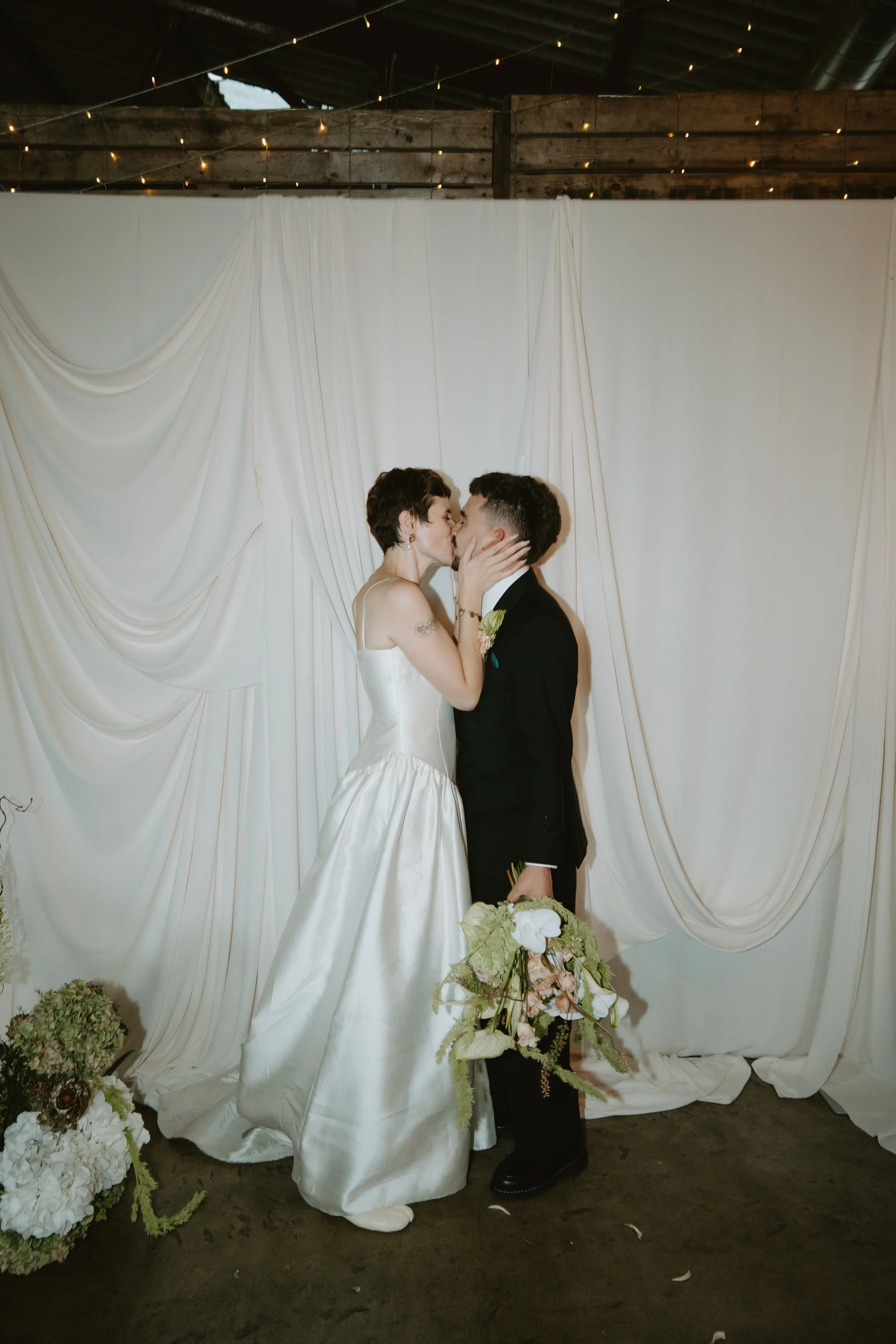 Orange Tree Weddings styling and décor creating a minimalist, romantic wedding backdrop with softly draped white fabric and warm festoon lighting, framing an intimate couple’s kiss for modern, editorial wedding photography.