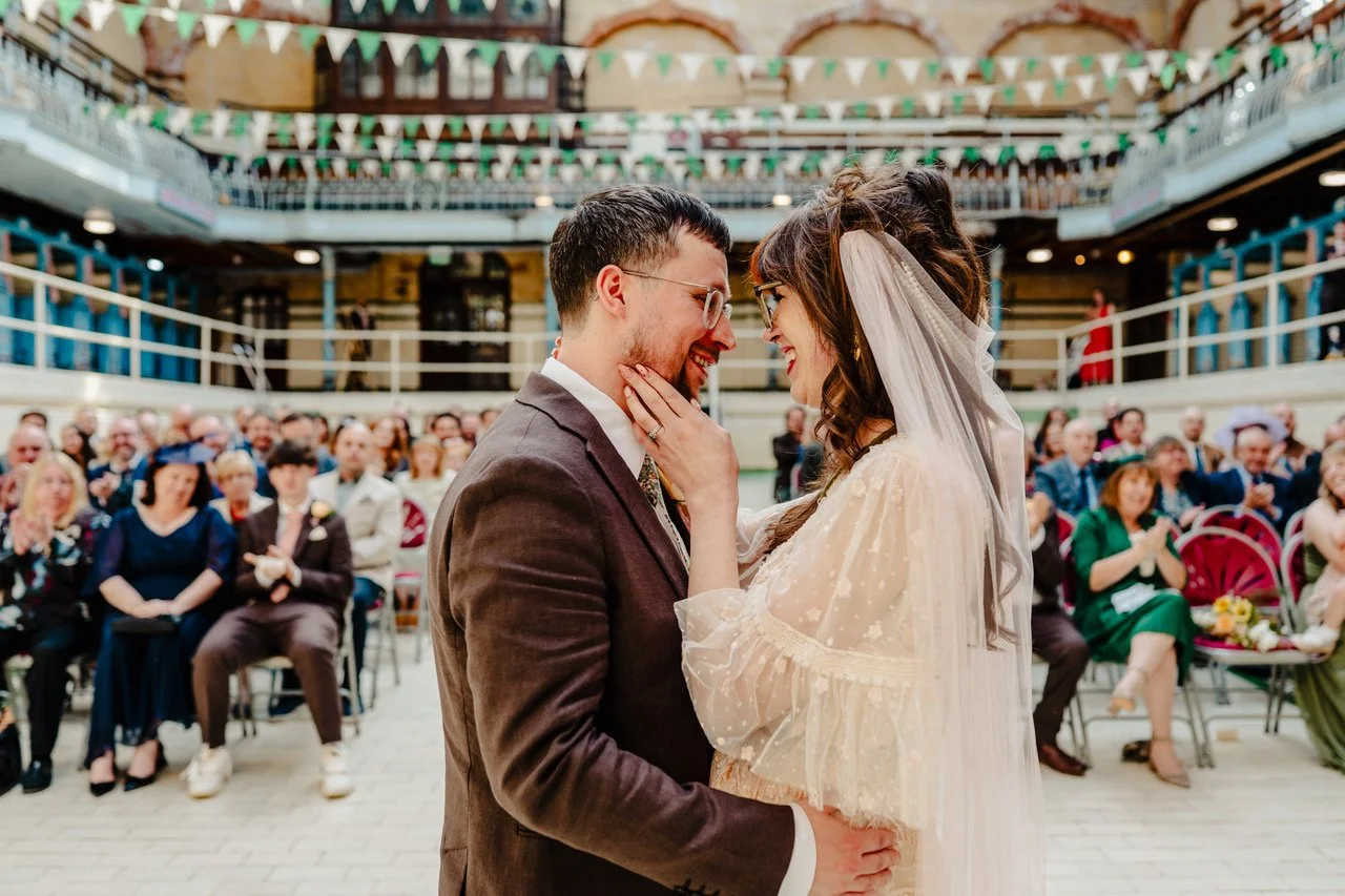 A couple stands wrapped in a tender embrace at the centre of their ceremony space with the bride smiling up at her partner as her hand rests on his cheek while guests seated behind them clap and beam beneath rows of green and white bunting that fill 