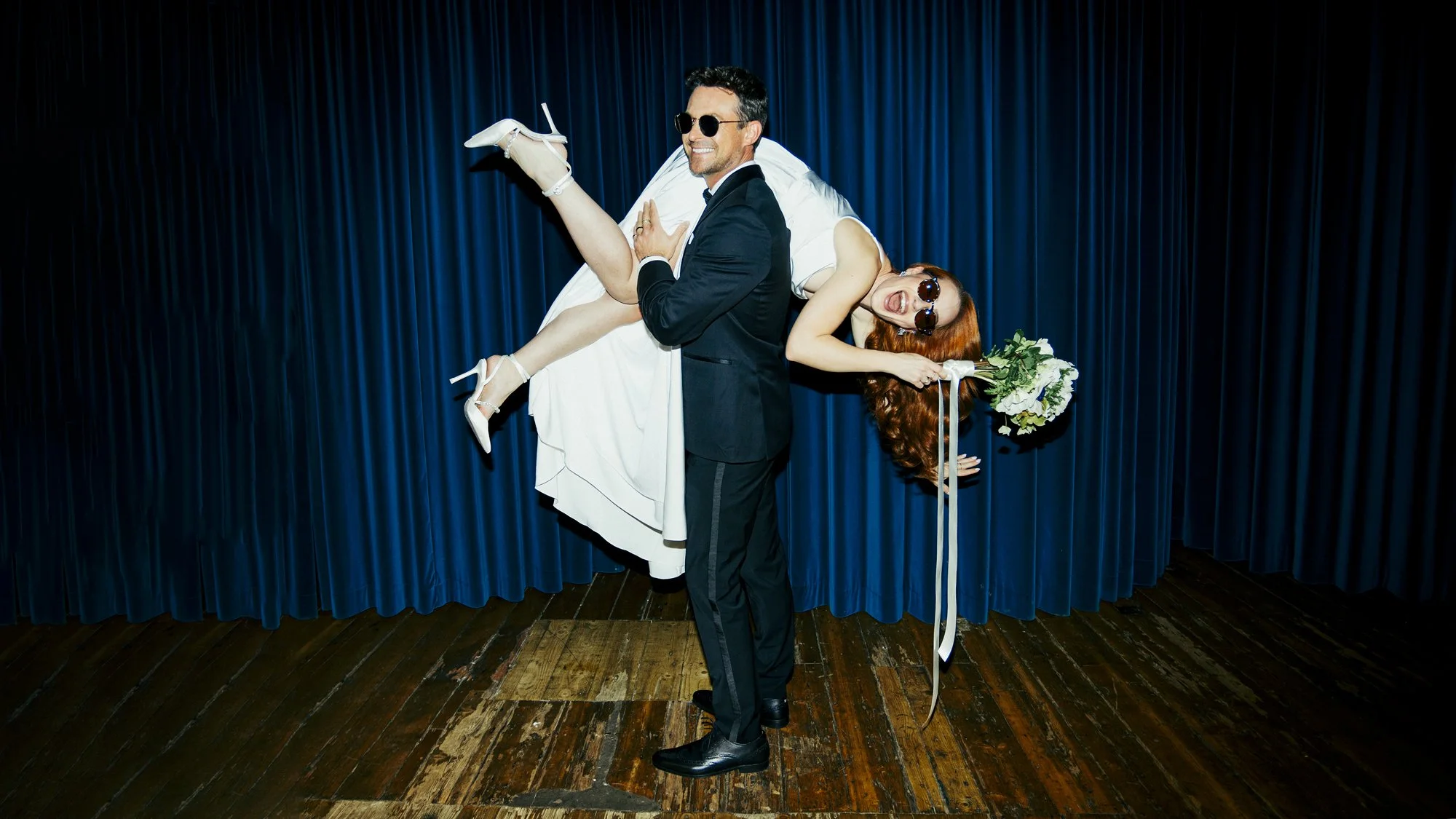 A playful and stylish wedding moment at Ministry Venues, featuring a newlywed couple striking a fun pose against rich blue velvet curtains, highlighting the venue’s bold character, theatrical backdrops and contemporary spaces that make it a standout 