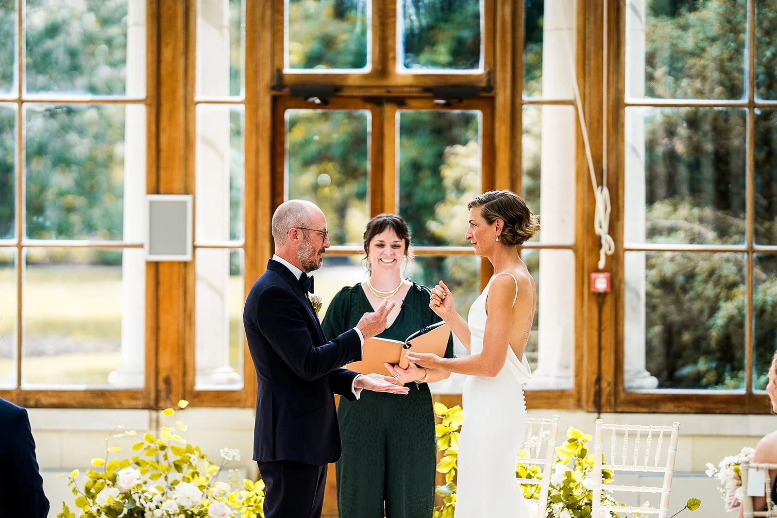 A joyful wedding ceremony led by a Cherry and Fern Celebrations, as a couple exchange vows and rings in a sunlit orangery surrounded by natural greenery and elegant floral arrangements. The moment captures the warmth and intimacy of a personalised we