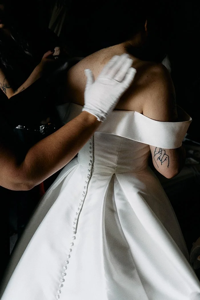 A bride wearing an off-the-shoulder satin wedding dress is gently adjusted by a gloved hand, highlighting the buttoned back, soft structure and intimate getting-ready moment captured by Elizabeth Royse Wedding Photography.