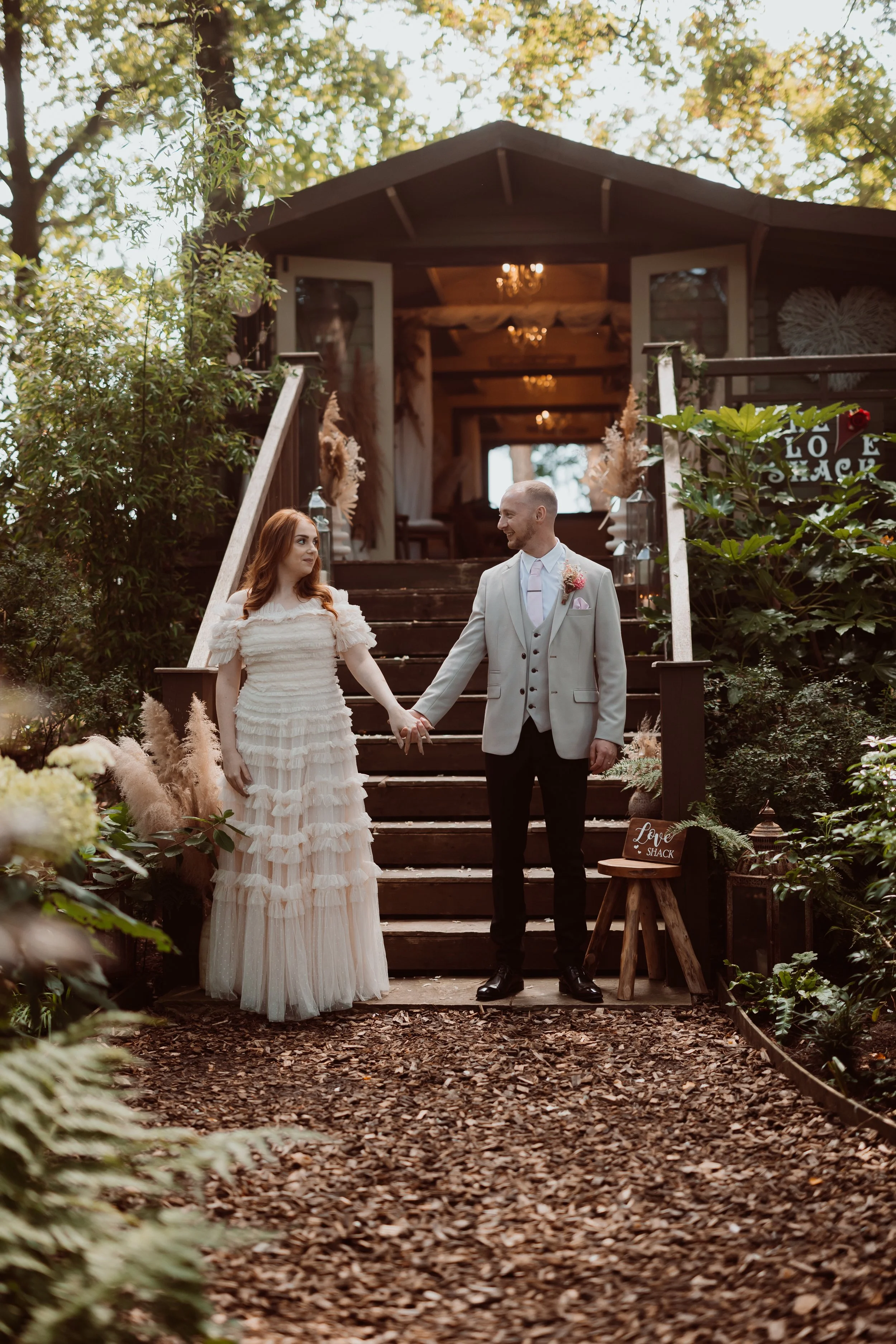 A romantic and storybook woodland moment captured by Joy Louise Photography, showing the couple standing hand in hand at the foot of a rustic timber staircase leading up to a beautifully decorated cabin. Surrounded by lush greenery, ferns and soft pa