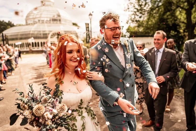 Joyful newlyweds celebrating outside Sefton Park Palm House in Liverpool, featuring the bride with radiant natural makeup by Glamorama Make-up Artist Liverpool. A vibrant and romantic wedding moment capturing fresh, dewy bridal beauty and modern boho