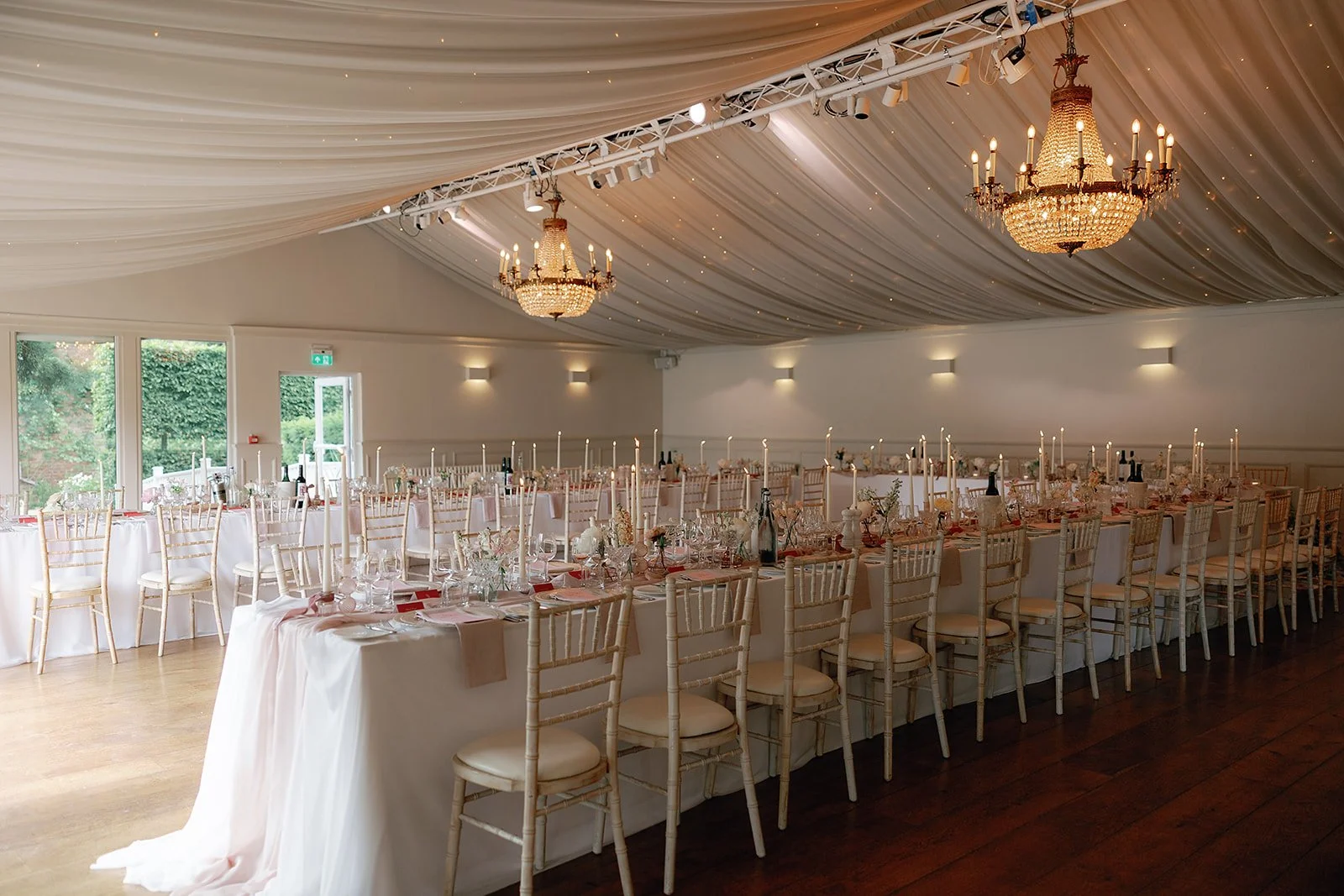 Soft draping stretches across the ceiling of the bright reception space while chandeliers cast a warm glow over the long tables that are dressed in white linens, pale pink runners, tall taper candles and delicate glassware with rows of elegant chairs