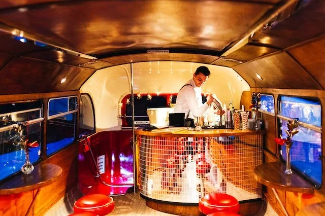 A bartender prepares cocktails inside the Deckerdence mobile bar’s retro double-decker bus lounge. The stylish curved bar, warm wood interior, and red leather stools create a vintage speakeasy atmosphere. Perfect for weddings, parties, and corporate 