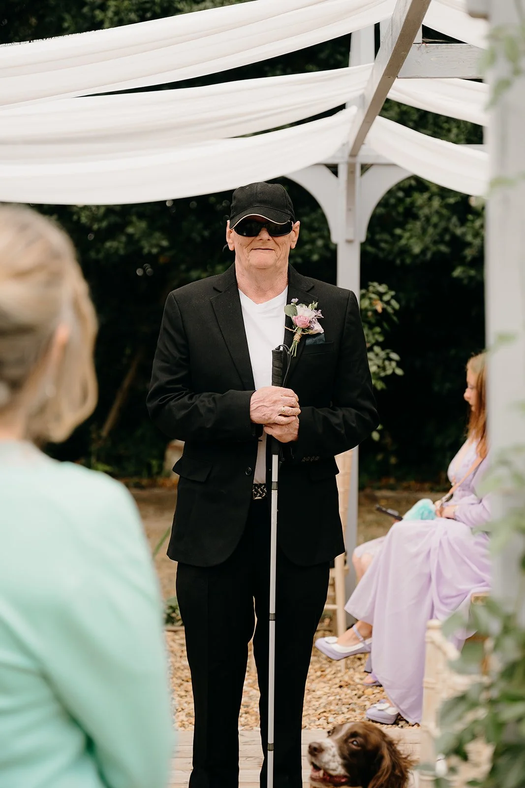 A very stylish groom is wearing a black suit with a floral buttonhole. He is blind and has black glasses on. He is holding a stick and waiting patiently for his bride to arrive. There are soft, whte drapes in the background and it is an outdoor cerem