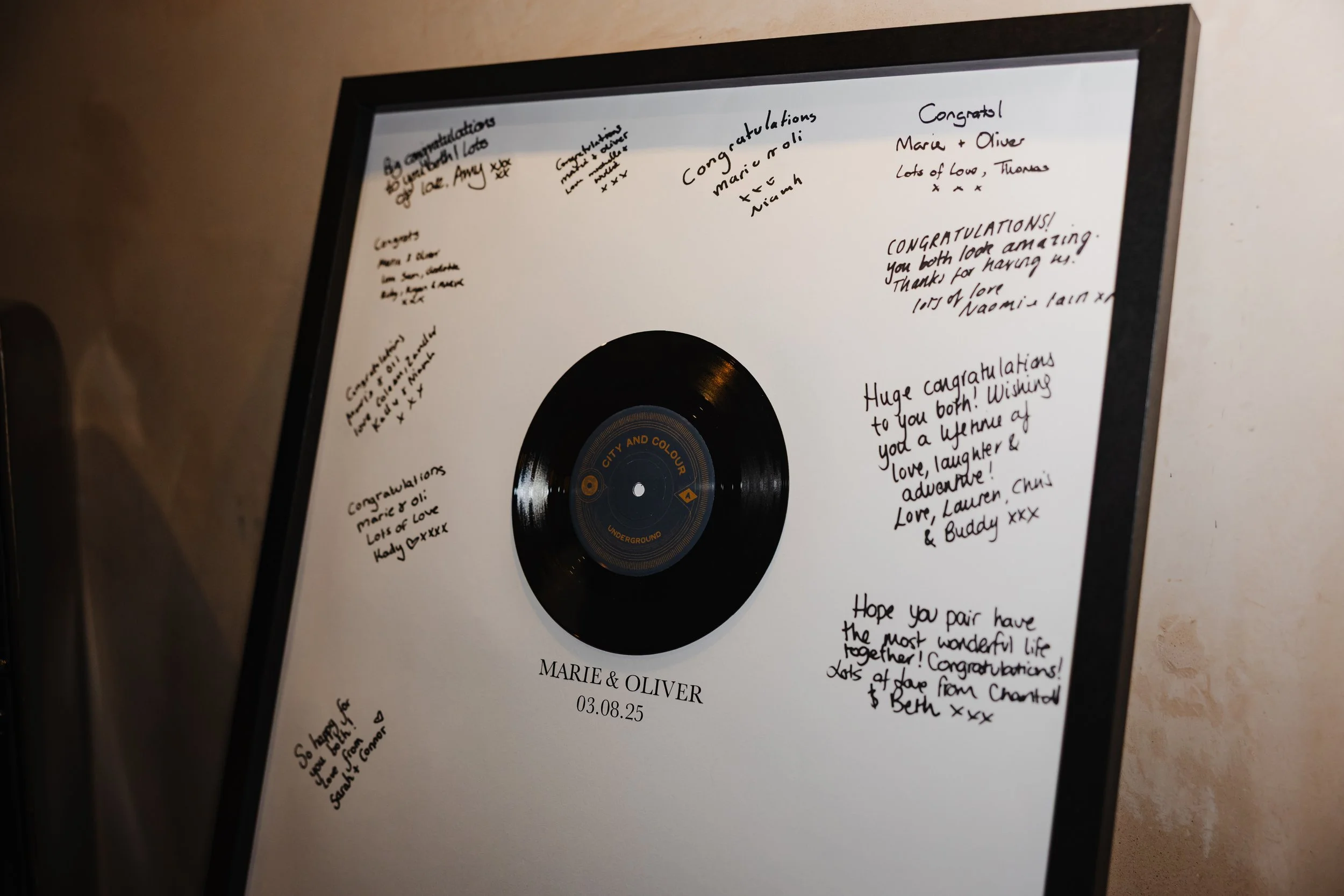 A black framed 'guestbook' styled display with a small vinyl record inset at the top, personalised with the couple’s names, “Marie & Oliver,” and their wedding date beneath. Guest have written well wishes in the space around it.