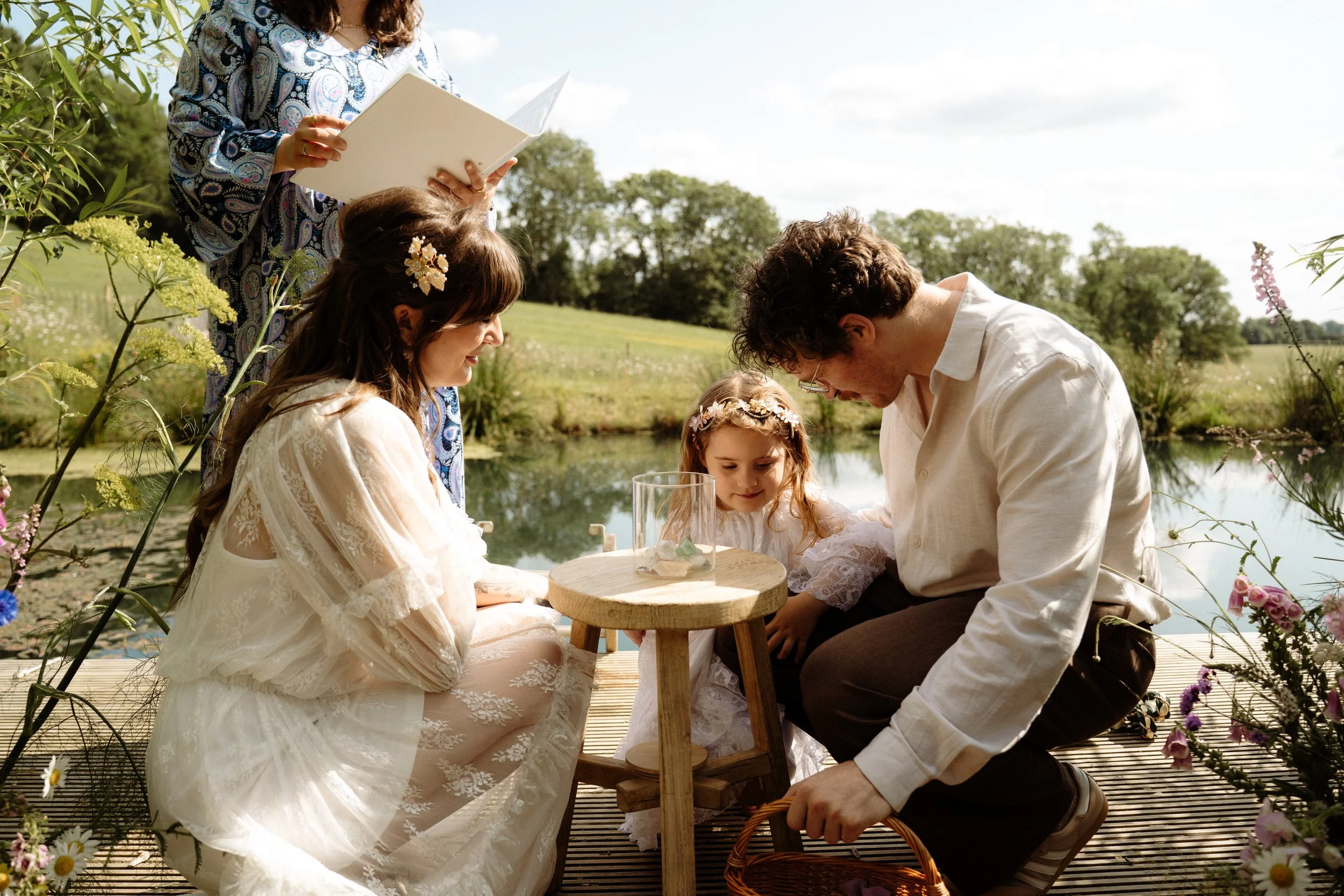 The couple kneels beside their young daughter by a tranquil pond, participating in a unity ritual involving a glass vase on a small wooden table - possibly a sand ceremony or symbolic keepsake ritual. Lauren The Spiritual Celebrant stands just behind