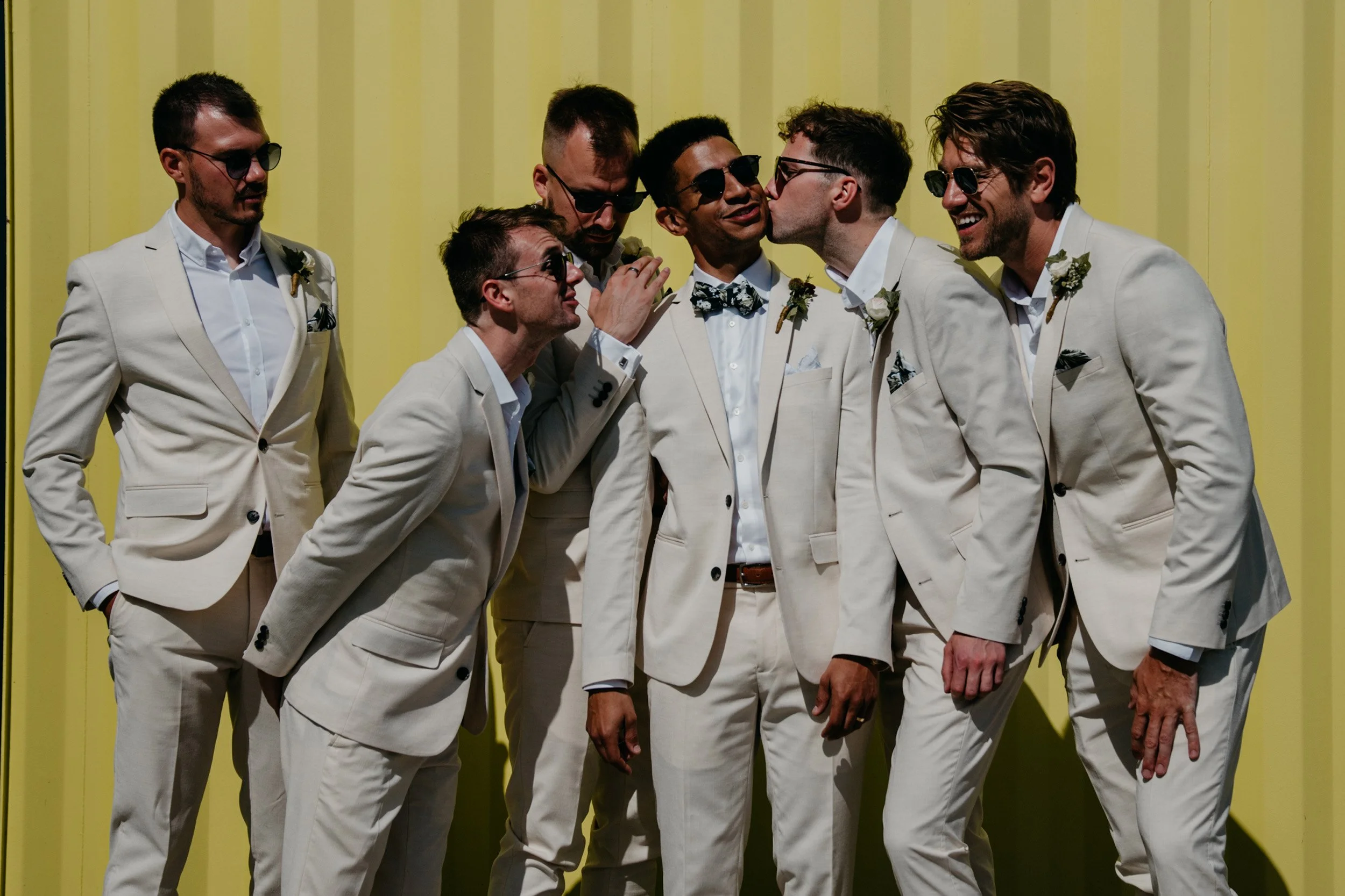 Six men stand together in front of a bright yellow wall wearing matching light cream suits with white shirts, floral bow ties and coordinated boutonnieres. Most of them wear sunglasses and lean in toward the groom at the centre, who smiles while one 