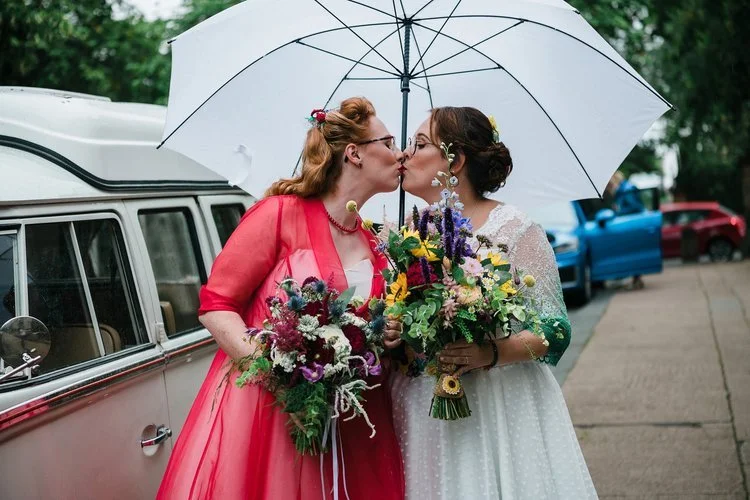 Colourful wedding bouquets by New Bud Floristry featuring vibrant seasonal blooms, textured foliage and wild garden accents held by two brides sharing a joyful moment under an umbrella. With bright expressive flowers and relaxed styling beside a vint