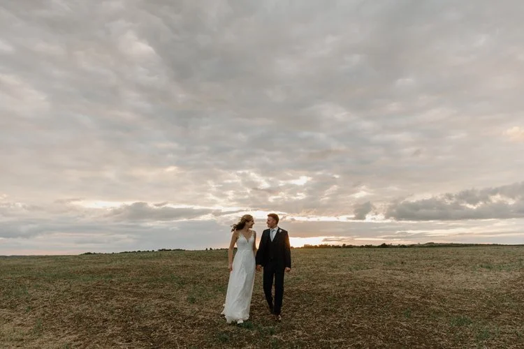 A peaceful and cinematic moment captured by YU Photography, showing the couple walking hand in hand across an open field beneath a dramatic evening sky. Soft golden light breaks through the clouds on the horizon, casting a warm glow over the landscap