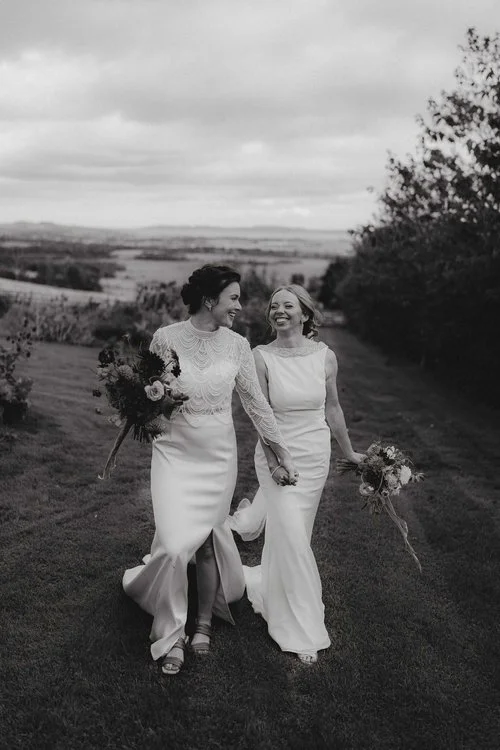 Two brides walk hand in hand across a windswept hillside, laughing together as their dresses flow behind them, beautifully captured by F.D Young Photography. The dramatic monochrome tones, open landscape and natural movement create an emotive, cinema