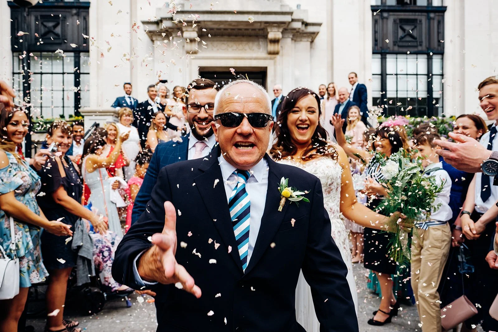 A man in a dark suit and striped tie walks toward the camera through a shower of confetti, wearing sunglasses and a boutonniere, while smiling guests line the steps of a grand building behind him. A woman in a white embellished dress stands just behi